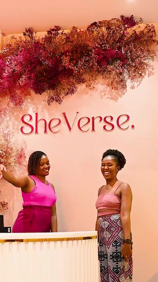 Two women laughing and pointing at the SheVerse logo wall in the studio reception